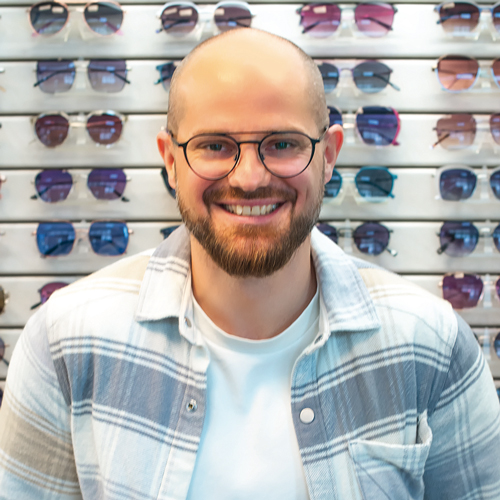 Mann mit Hemd und einer Brille steht vor einer Wand mit Sonnenbrillen.